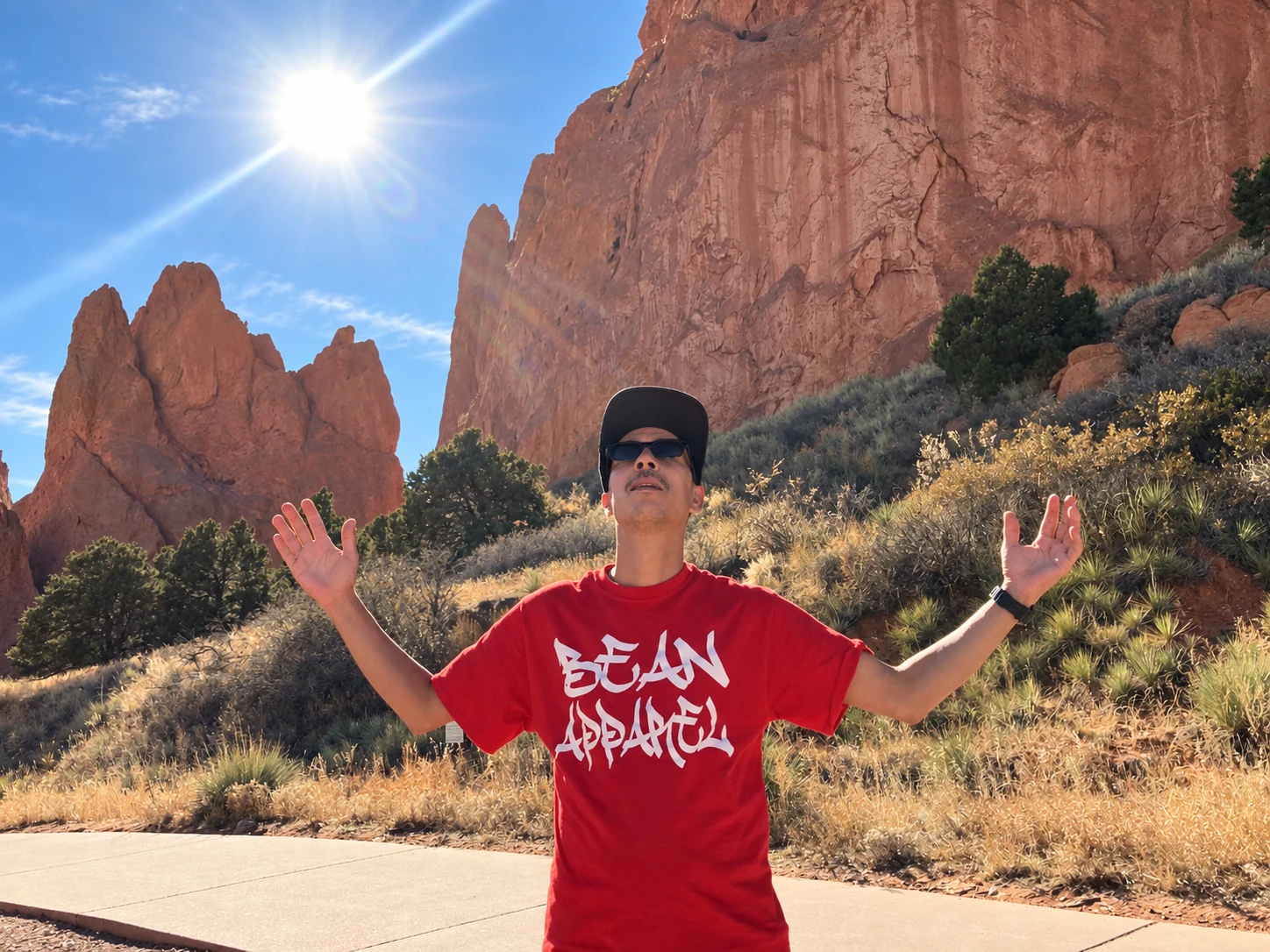 Man wearing a red Bean Apparel shirt with arms outstretched beneath sunlit rock formations
